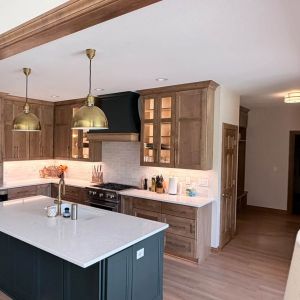 Open concept kitchen remodel featuring dark center island, brass pendant lights, warm wood cabinets, and custom range hood.   
