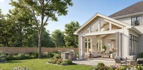 A wide photograph of a luxury home addition with open folding glass doors revealing a modern living room, looking out onto a spacious stone patio, outdoor grill station, and fenced backyard with mature trees.